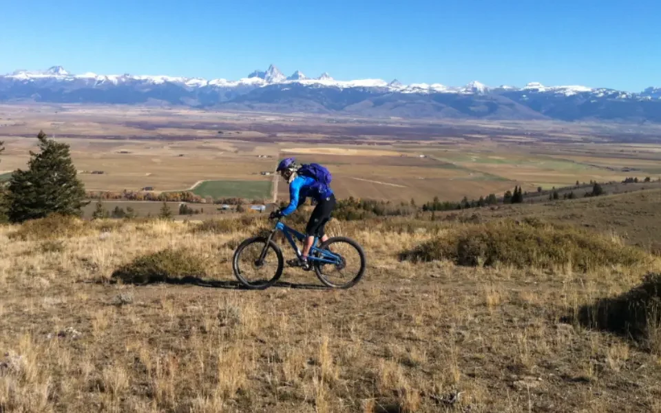 Mountain biker descending a high ridge trail above a sweeping Jackson Hole valley with snow-capped Teton peaks on the horizon