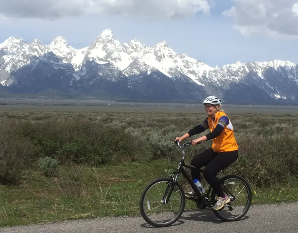 Smiling woman in an orange vest riding a bike on a paved path with the Grand Teton Range filling the skyline